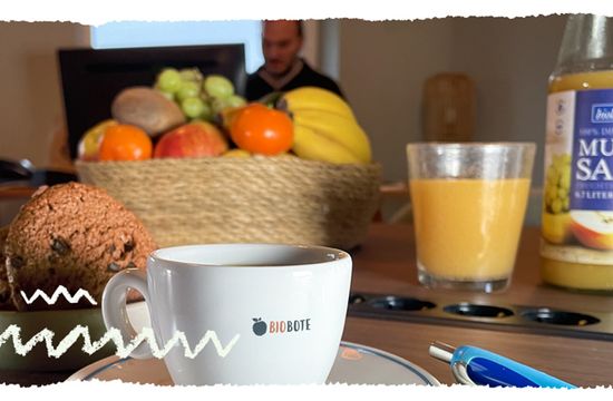 Büro Bepsrechungstisch mit Kaffe, Keksen, friscger Obstkorb, Saft. Im Hintergrund arbeitet jemand am Schreibtisch