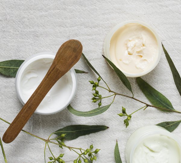 KI generiert: Cremetiegel mit Holzspatel und grünen Zweigen auf hellem Stoff.