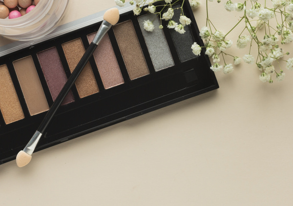 KI generiert: Lidschatten-Palette und Blüten auf beigem Hintergrund, begleitet von einem Applikator und Rouge.