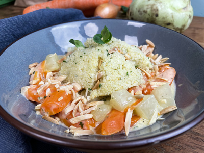 KI generiert: Das Bild zeigt ein Gericht mit Couscous, das mit geschnittenem Gemüse wie Karotten und Kohlrabi sowie gerösteten Mandelsplittern in einer cremigen Sauce angerichtet ist. Im Hintergrund sind frische Zutaten wie Karotten und Kohlrabi zu sehen.