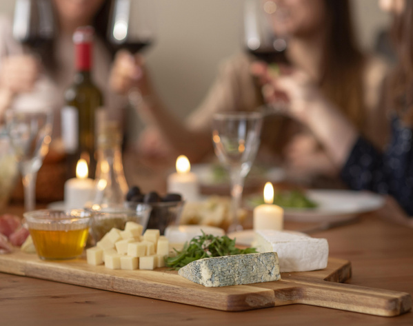 KI generiert: Verschiedene Käsesorten auf einem Holzbrett, Menschen im Hintergrund stoßen mit Rotwein an.