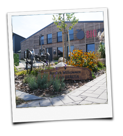 KI generiert: Außenaufnahme eines Gebäudes mit Blumenbeet und Schild: "Herzlich Willkommen bei den BIOBOTEN".