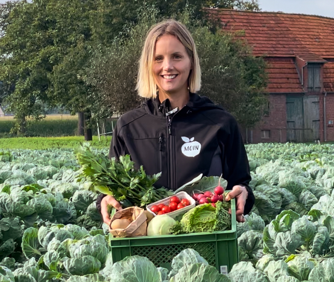 KI generiert: Eine Person hält eine Kiste mit frischem Gemüse in einem Feld. Jacke mit "MOIN"-Logo.