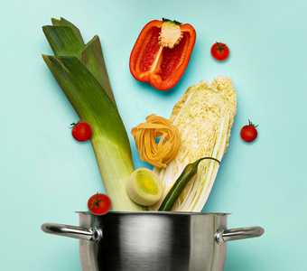 Ein Kochtopf mit Lauch, Paprika, Tomaten, Nudeln und Chinakohl auf hellblauem Hintergrund.