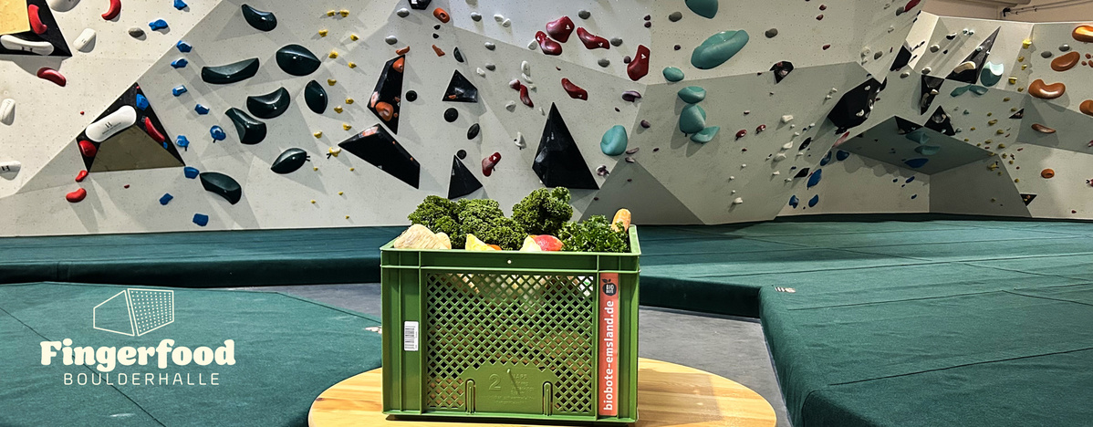 KI generiert: Eine Kiste Gemüse vor einer Boulderwand in einer Halle. Text: "Fingerfood Boulderhalle".