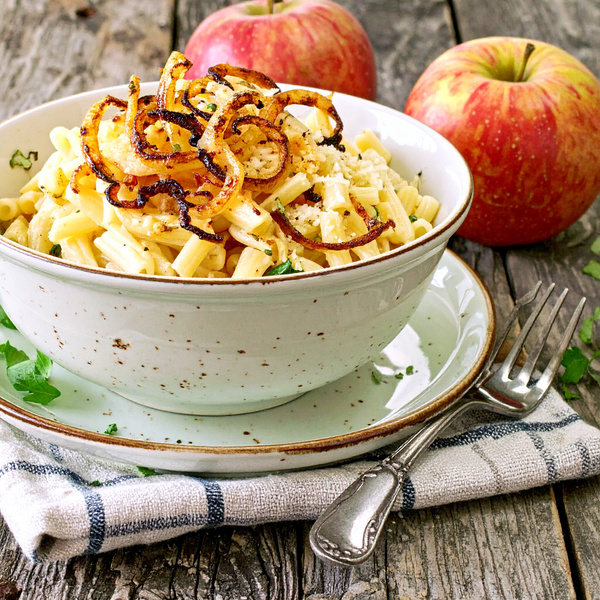 KI generiert: Eine Schüssel Pasta mit gebratenen Zwiebeln, umgeben von zwei Äpfeln auf einem rustikalen Tisch.