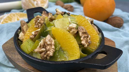 KI generiert: Das Bild zeigt einen Salat, der aus Orangenscheiben, Walnüssen und Lauch besteht, serviert in einer kleinen schwarzen Pfanne. Im Hintergrund sind Orangen und Walnüsse zu sehen.