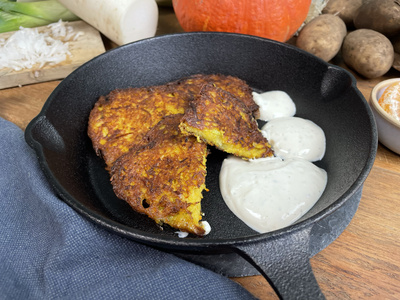 KI generiert: Das Bild zeigt zwei knusprige Kartoffelpuffer in einer schwarzen Pfanne, serviert mit einem Klecks Rahm. Im Hintergrund sind verschiedene Gemüse wie Kürbis und Kartoffeln zu sehen.