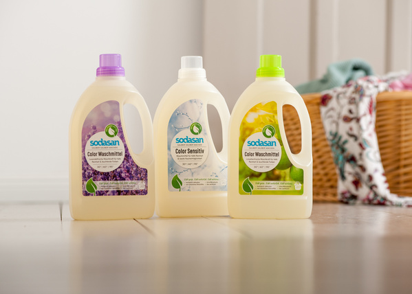 KI generiert: Drei Flaschen Sodasan Waschmittel auf dem Boden vor einem Wäschekorb. Text: "Color Waschmittel", "Color Sensitiv".