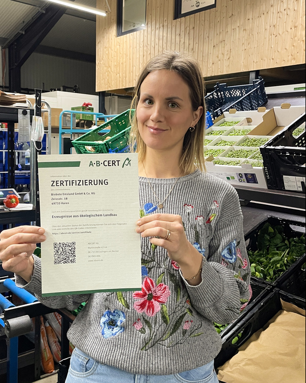 Lisa hält ein Zertifikat für ökologischen Landbau in einem Raum mit frischem Gemüse. Im Hintergrund sind Kisten mit Gemüse zu sehen.