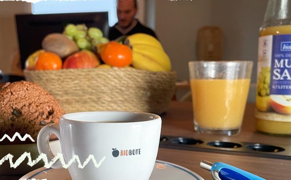 Büro: Mann sitzt am Computer. Im Vordergrund auf dem Besprechunstisch steht eine Schale frisches Bio-Obst, Orangensaft, Kekse und eine Tasse Kaffee