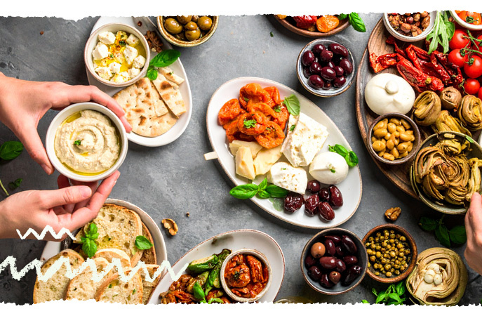 KI generiert: Verschiedene mediterrane Gerichte auf Tellern, darunter Käse, Oliven und Antipasti. Hände greifen nach Schalen.