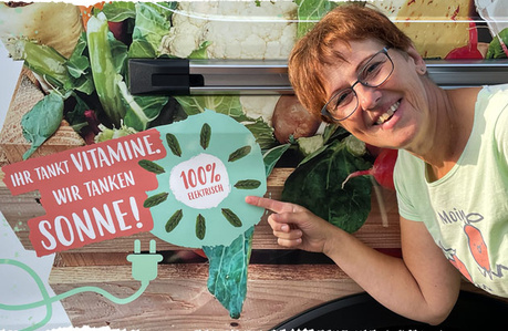 Eine Frau zeigt auf ein 100% elektrisches Fahrzeug mit Gemüse-Hintergrund. Text: "IHR TANKT VITAMINE. WIR TANKEN SONNE!"