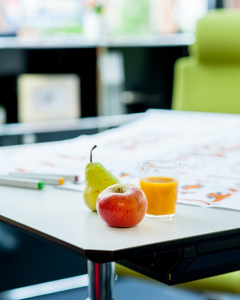 Das Bild zeigt einen Schreibtisch mit einem Apfel, einer Birne und einem Glas Orangensaft. Im Hintergrund sind verschwommene Büromöbel zu sehen.