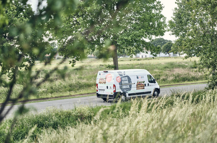 KI generiert: Ein weißer Lieferwagen fährt auf einer Straße, umgeben von grünen Bäumen. Text: „FRISCH. EHRLICH.“