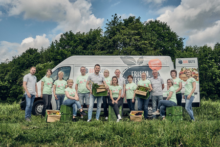 Eine Gruppe Menschen posiert mit Gemüsekisten vor einem Lieferwagen auf einer Wiese. Text: "BIObote".