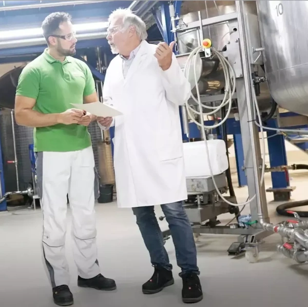 KI generiert: Zwei Männer diskutieren in einem industriellen Umfeld. Einer trägt einen Laborkittel.