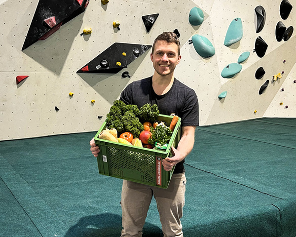 KI generiert: Ein Mann hält eine grüne Kiste mit Gemüse vor einer Kletterwand. Text: "die-gemüsegärtner.de".