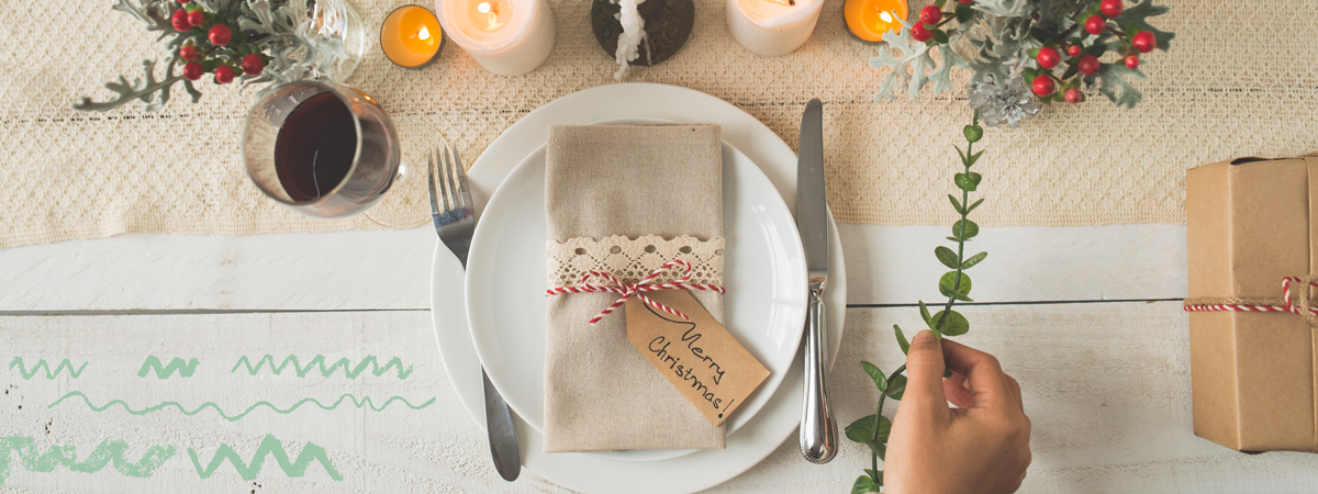 KI generiert: Das Bild zeigt einen weihnachtlich dekorierten Tisch mit einem eingedeckten Teller, Besteck und einem mit einem "Merry Christmas"-Schild verzierten Serviettenpaket. Im Hintergrund sind Kerzen, weihnachtliche Dekorationen und ein Geschenk zu sehen.