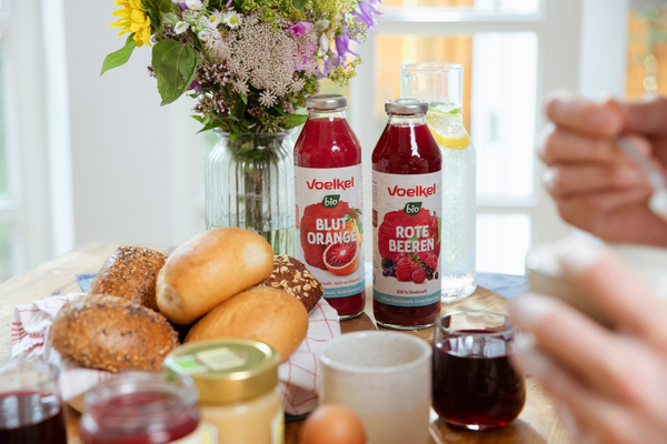 KI generiert: Frühstückstisch mit Voelkel Bio-Säften "Blutorange" und "Rote Beeren", Blumen und Brötchen.