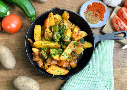 KI generiert: Das Bild zeigt eine Pfanne mit gebratenen Kartoffeln und Gemüse, garniert mit frischen Kräutern. Um die Pfanne herum liegen frische Zutaten wie Tomaten, Zucchini und Gewürze.
