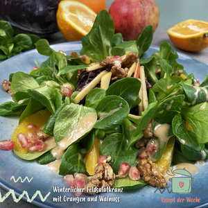 Das Bild zeigt einen winterlichen Feldsalatkranz mit Orangen, Walnüssen, Apfelstreifen und Granatapfelkernen auf einem Teller, umgeben von ganzen Orangen und einem Rotkohl im Hintergrund. Der Salat ist als "Rezept der Woche" gekennzeichnet.
