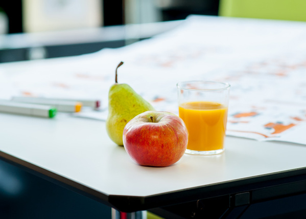 Das Bild zeigt einen Schreibtisch mit einem Apfel, einer Birne und einem Glas Orangensaft. Im Hintergrund sind verschwommene Büromöbel zu sehen.