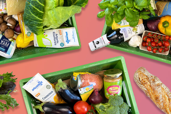 Grüne Kisten mit frischem Gemüse, Brot und anderen Lebensmitteln vor rosa Hintergrund.